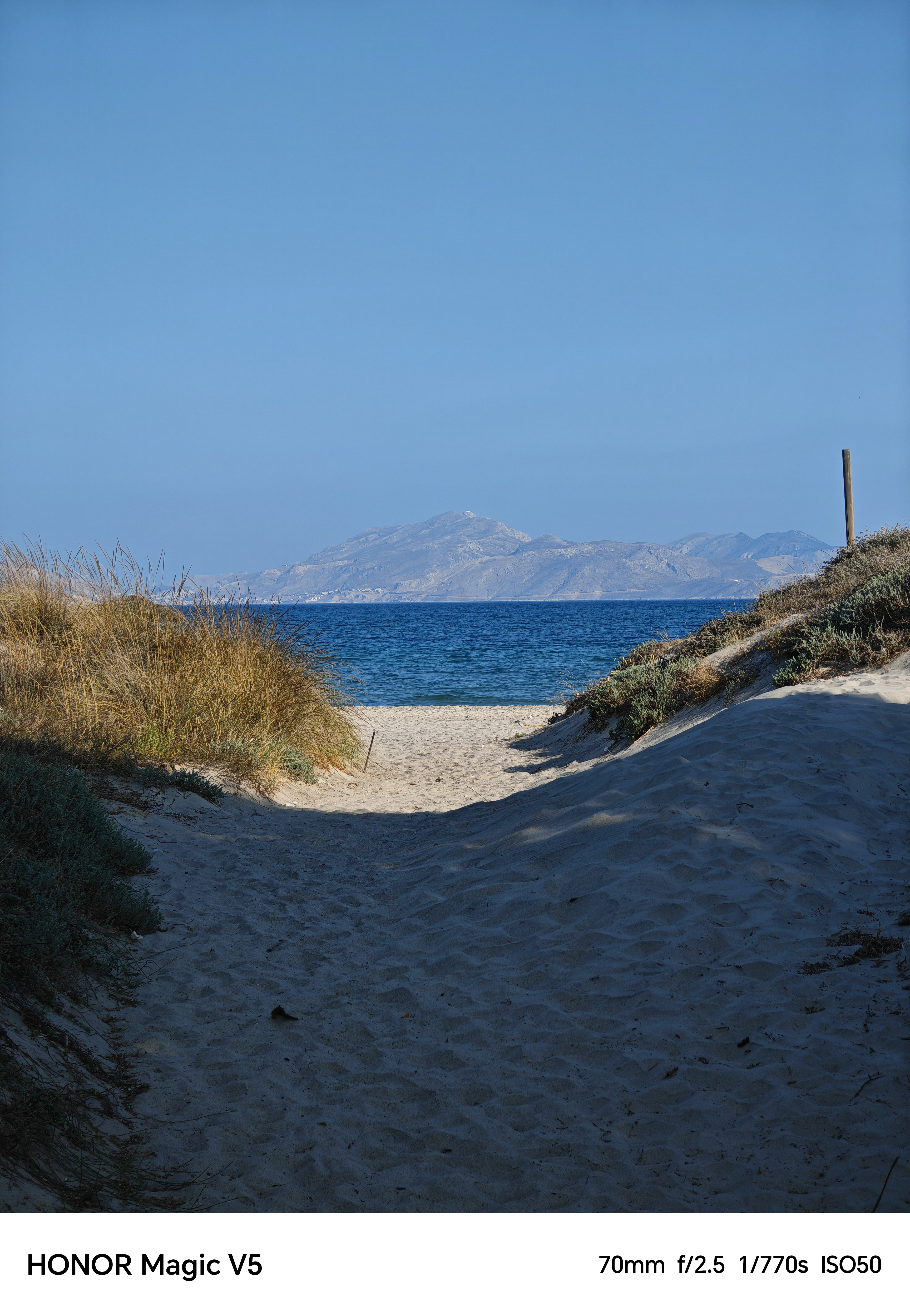 A sandy path leading down to the ocean with blue skies and mountains in the background