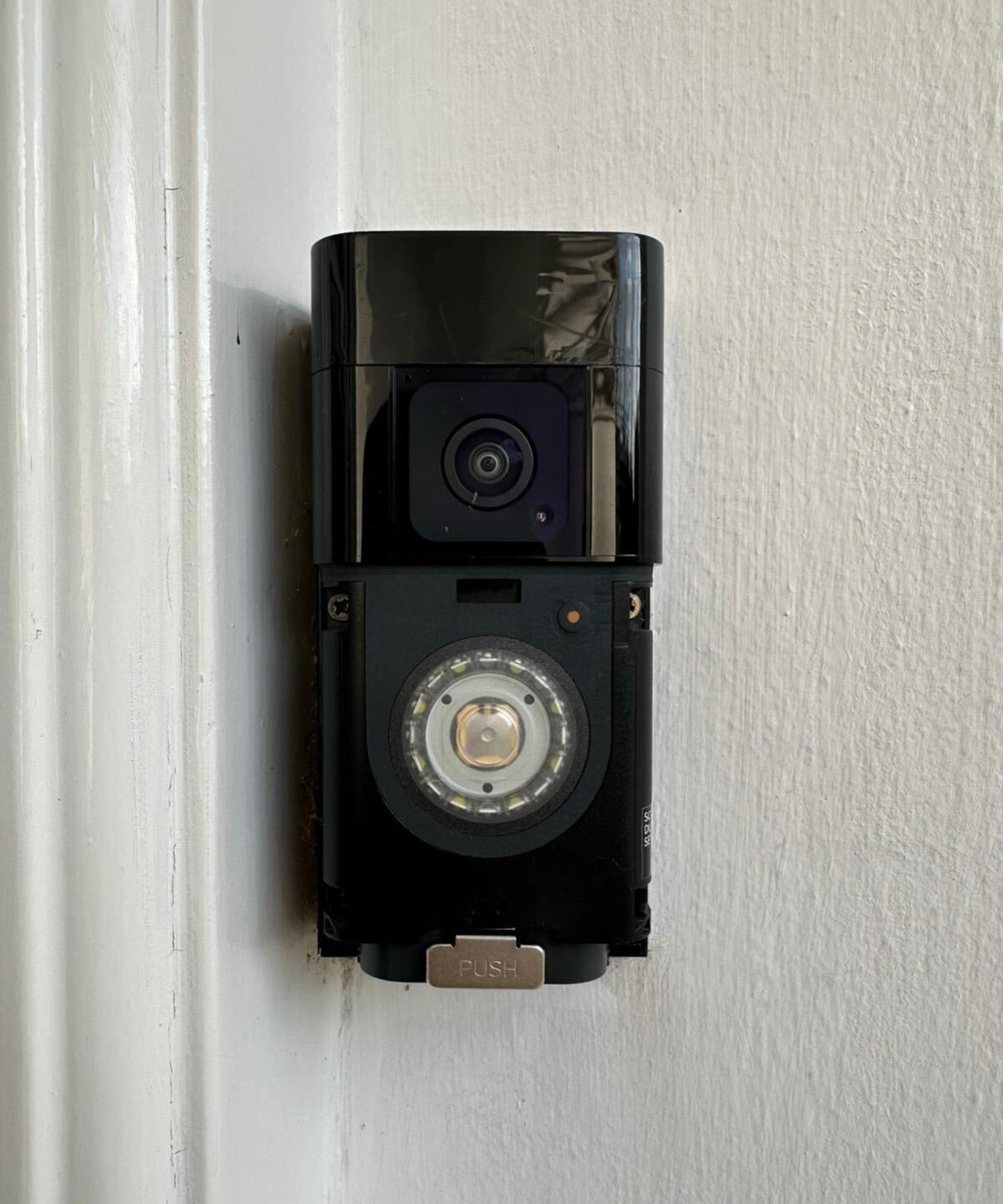 A black Ring Battery Doorbell Pro without a faceplate mounted on a painted white wall with a painted white architrave to the left.