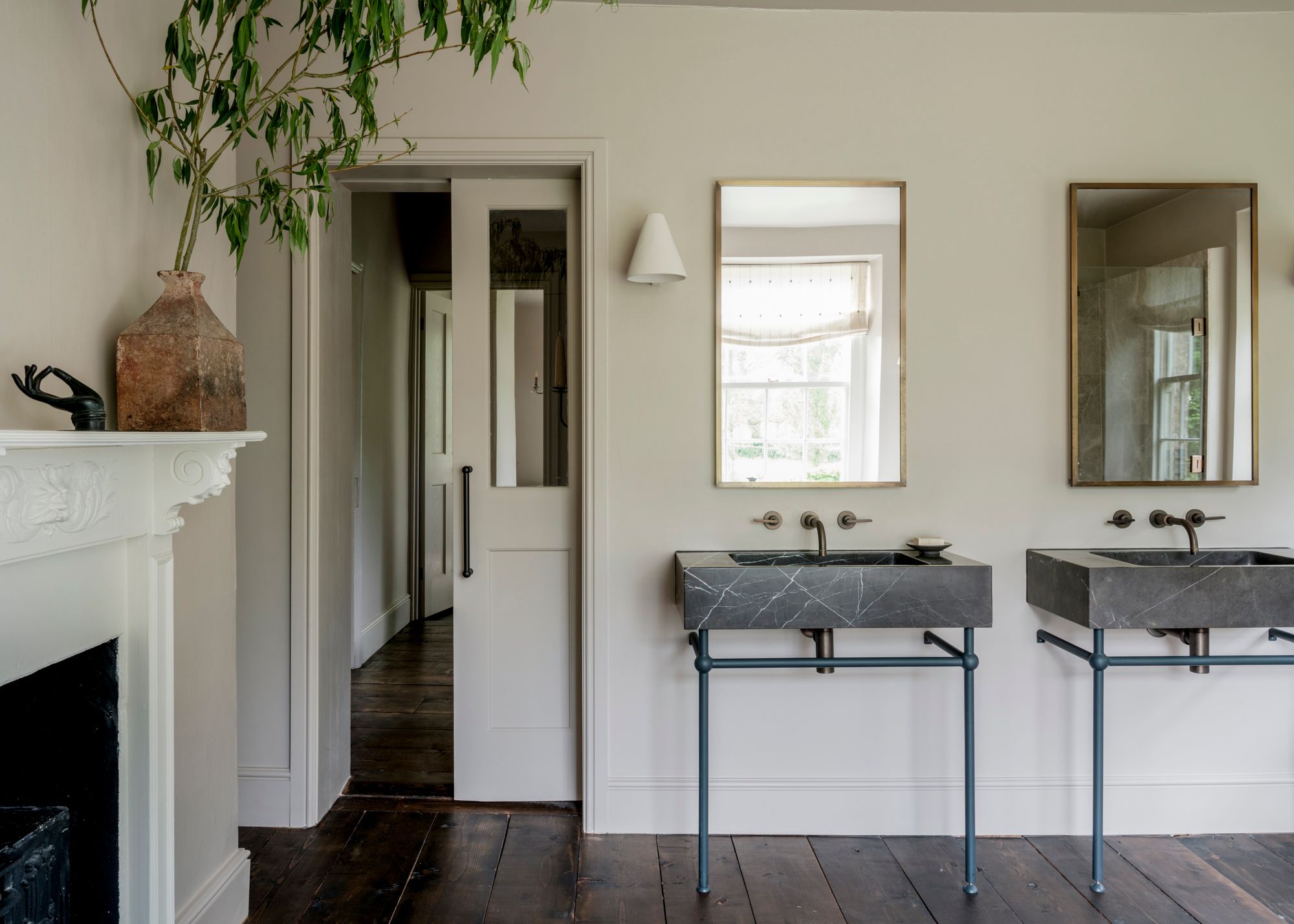 A large bathroom in a historic home with neutral color-drenched walls, dark wood flooring, double sinks with modern marble basins, rectangular mirrors on the wall, and a fireplace in the corner.