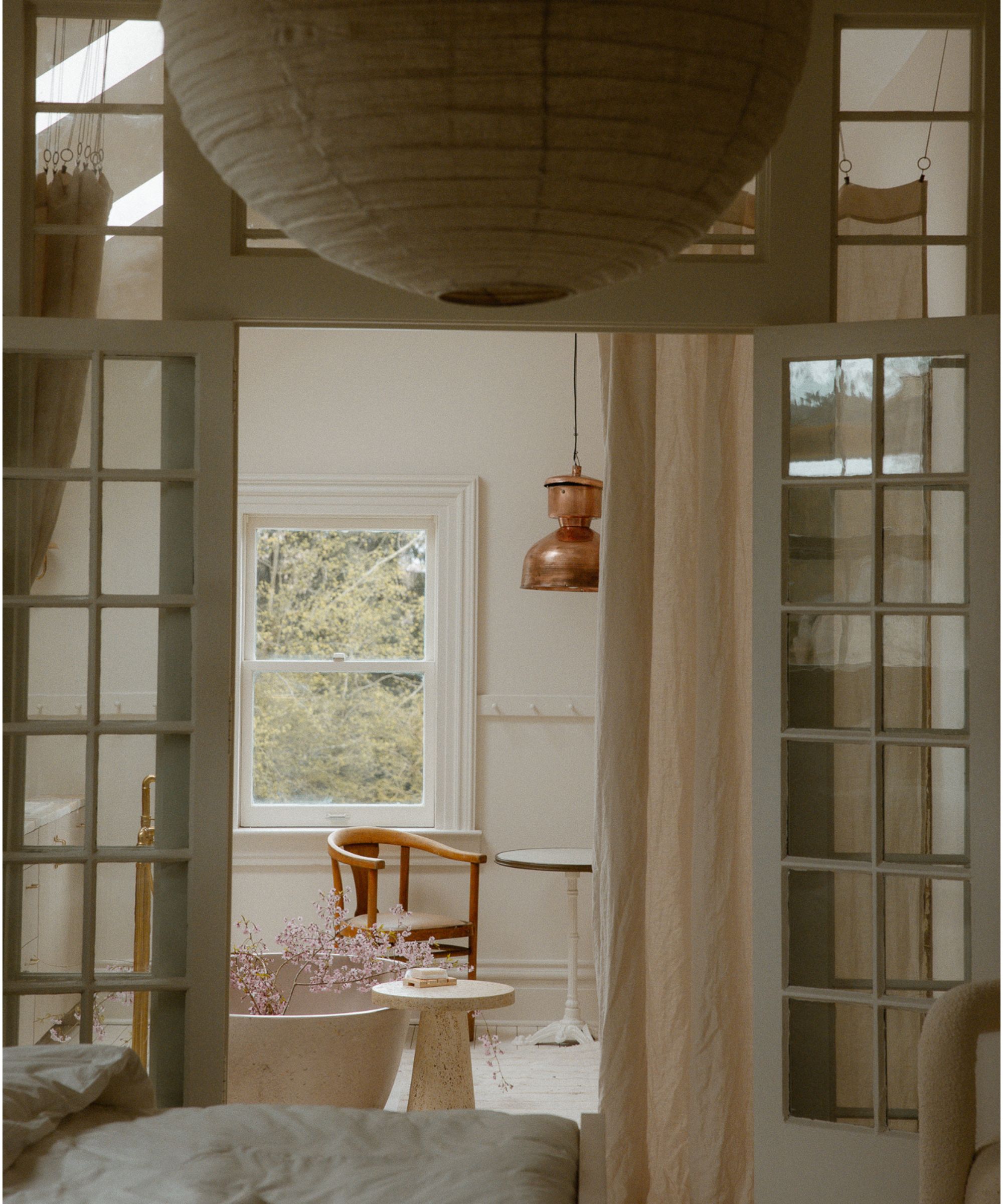 Bedroom with white walls, window with a wooden chair and round table beneath, glass doors leading to sleep space with linen drapes and a bed