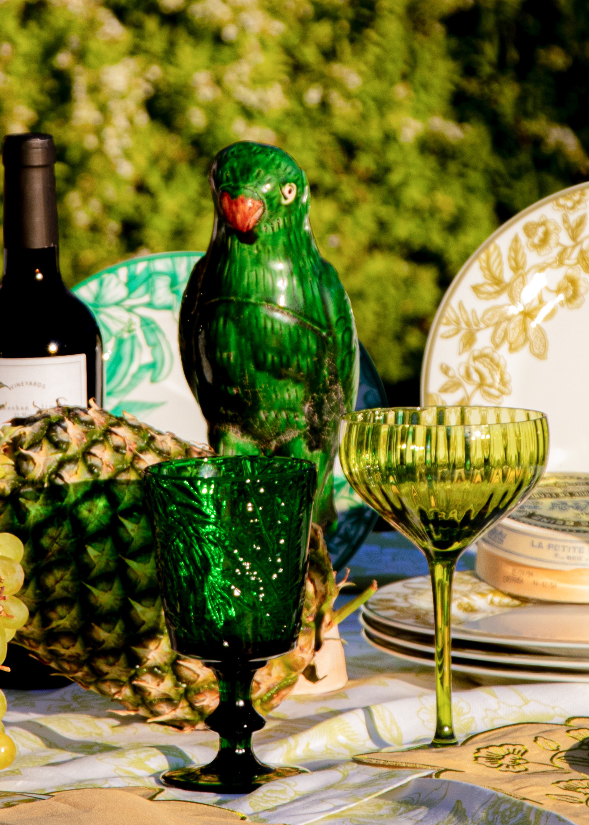 A close up of a tablescape with green glasses, fruit, and a parrot figuring as the centerpiece
