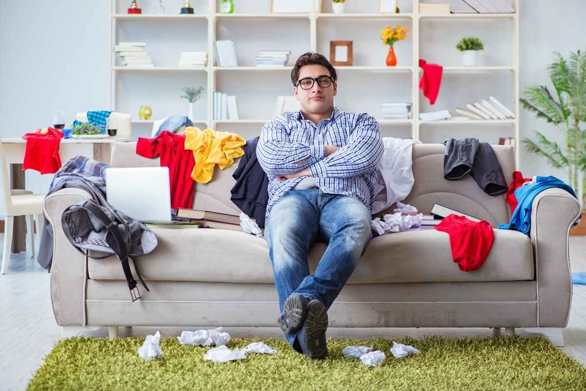 Man sitting on couch in a messy room.