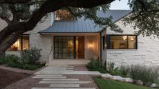 exterior of home with black sloped roof, white stone, garden beds, and staggered pavers in gravel path, with large tree reaching over