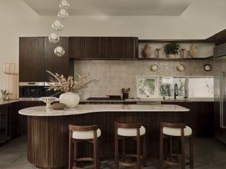 A modern kitchen with brown cabinets, a curved marble island, and boucle upholstered bar stools