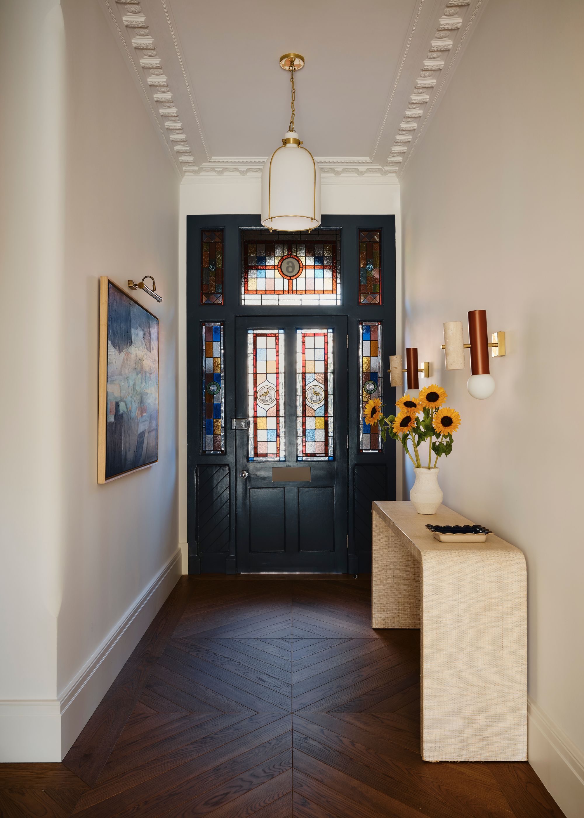 modern entryway with dark wooden floor, white walls high ceiling, light wood console table and traditional front door with colored glass panels