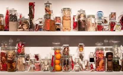 Two shelves featuring a range of objects in glass containers, including baby dolls, oranges, acorns. 