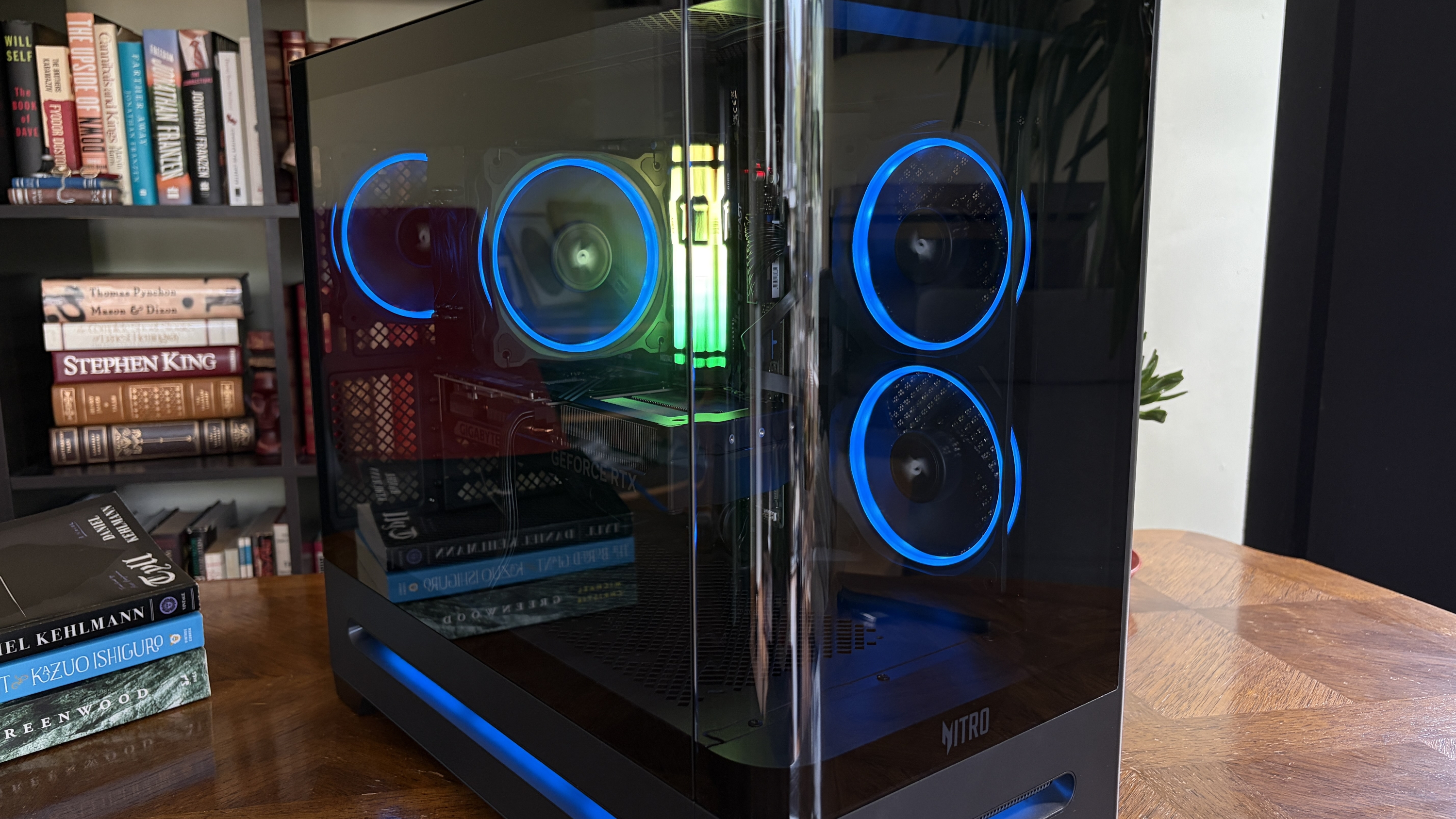 A black Nitro 65 gaming computer tower with a glass side panel and blue LED lights inside, displayed on a wooden table with books in the background.