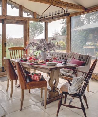 Wood beamed orangery with mismatched wooden dining table and chairs, with a horizontal candelabra suspended from above