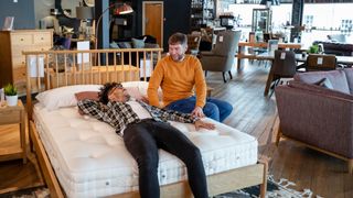 A man lying on a mattress testing it in a shop and his friend sat on the edge of the bed smiling