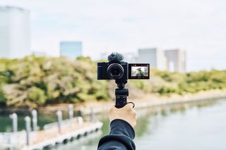 Sony ZV-1 II camera in the hand in vlogging mode with tree-fronted river blurred in background