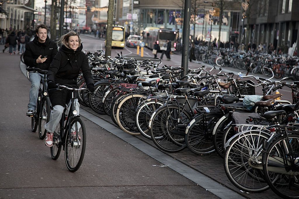Two people cycle next to a row of parked bikes