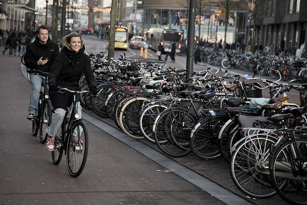 Two people cycle next to a row of parked bikes
