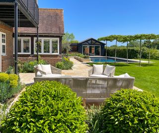 View of a sunny stone patio seating area near a house with a pool in the background surrounded by trees and other planting