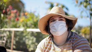 Sarah, the patient in the clinical trial, in her garden; she's wearing a floppy hat and surgical mask and appears to be smiling