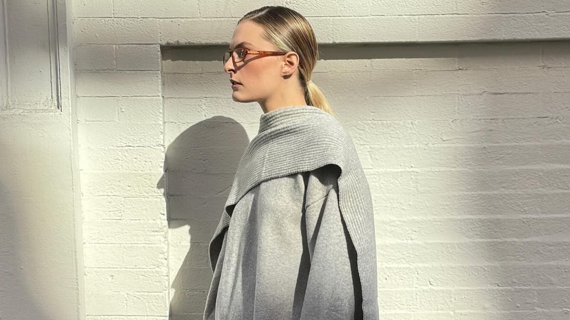 Influencer @christietyler poses outside in front of a beige brick wall wearing a grey cape jumper with a tonal grey skirt, thick black tights and black heels.