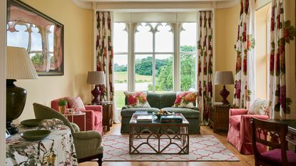 Living room with wooden floor and gothic window with views to the garden. Red and green upholstered chairs and sofa and floral curtains and cushions.