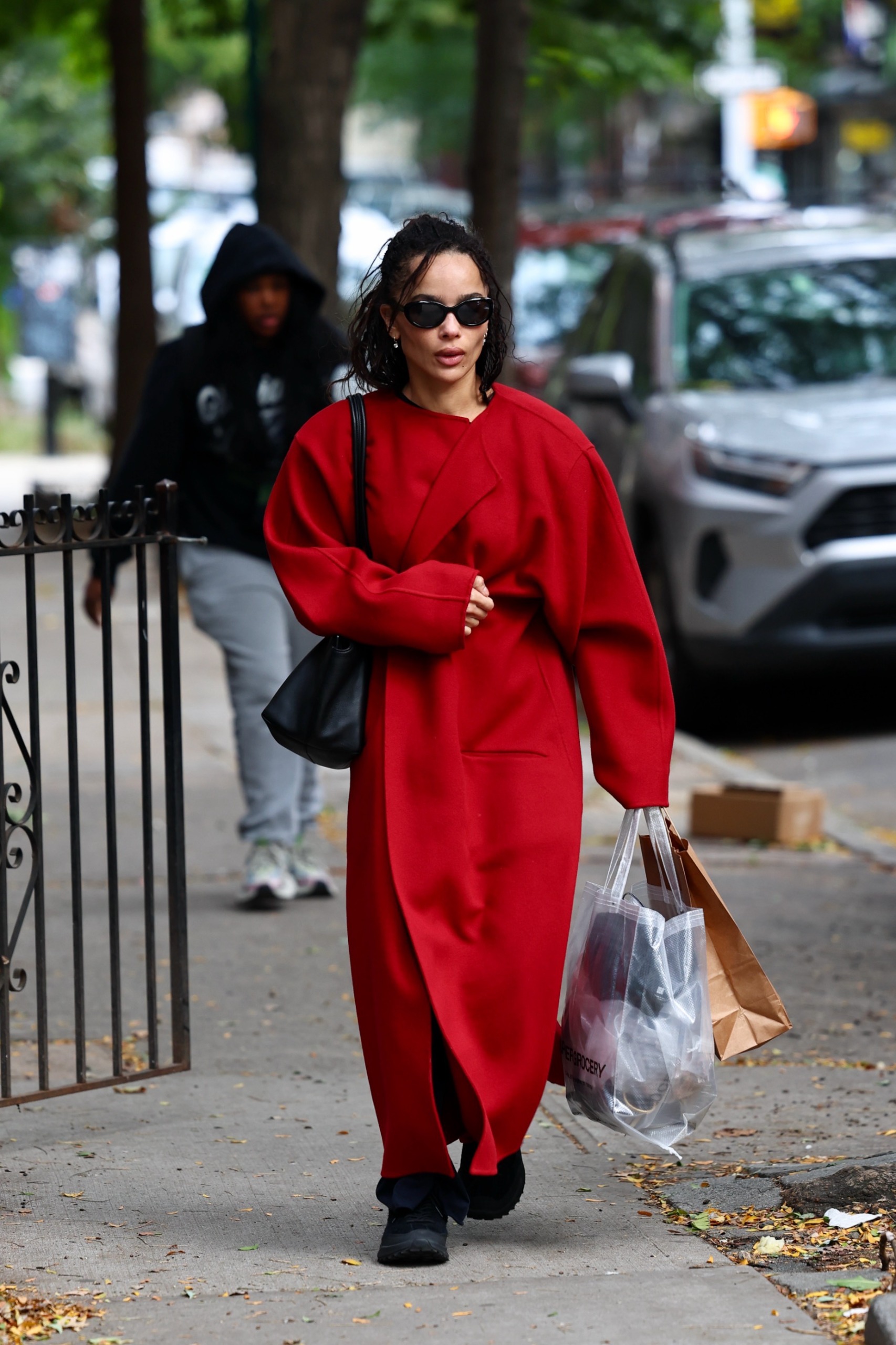 Zoe Kravitz walks down the street in New York wearing a red collarless coat with black trainers, a black leather bag and black sunglasses. She holds a transparent shopping bag and a brown paper bag.