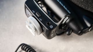 a black shotgun microphone by joby called the wavo pro is photographed by itself against a blue background, showing its muff, dead cat, attachment foot, and connection ports