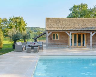 A large pool with a patio, a wicker dining area and a wooden pool house
