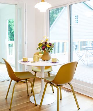 A white dining room with a white circular table with flowers and yellow tableware, two light brown plastic chairs, a pendant light over the table, and glass windows looking onto a house and balcony