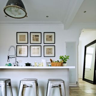 White kitchen, pale grey island unit, metallic bar stools, framed prints on feature wall, pendant light, natural wood flooring