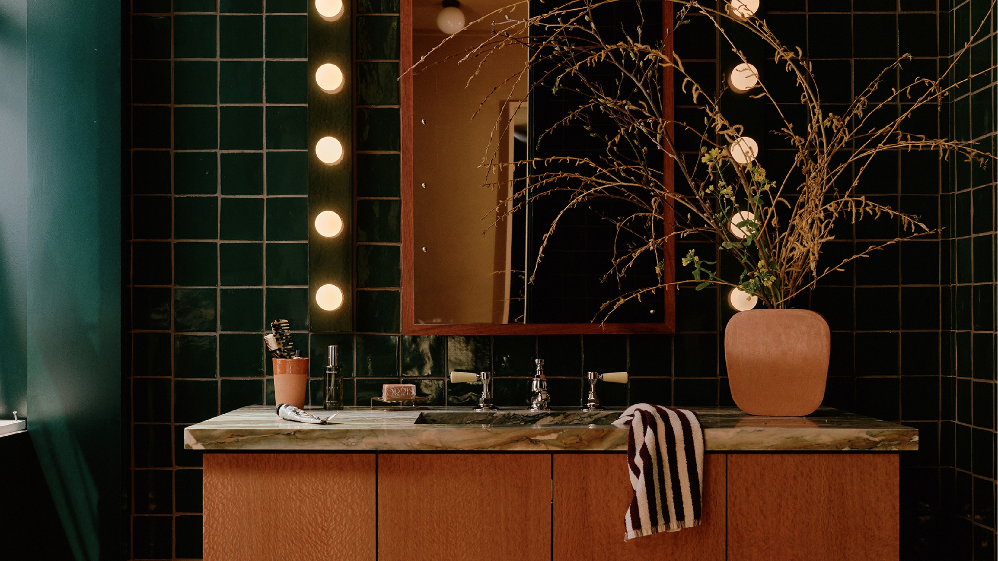 a green tiled and painted bathroom with hollywood vanity lighting and a marble and wood vanity, with a striped washcloth