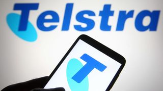 The Telstra logo on a smartphone with the company's name on the wall in the background