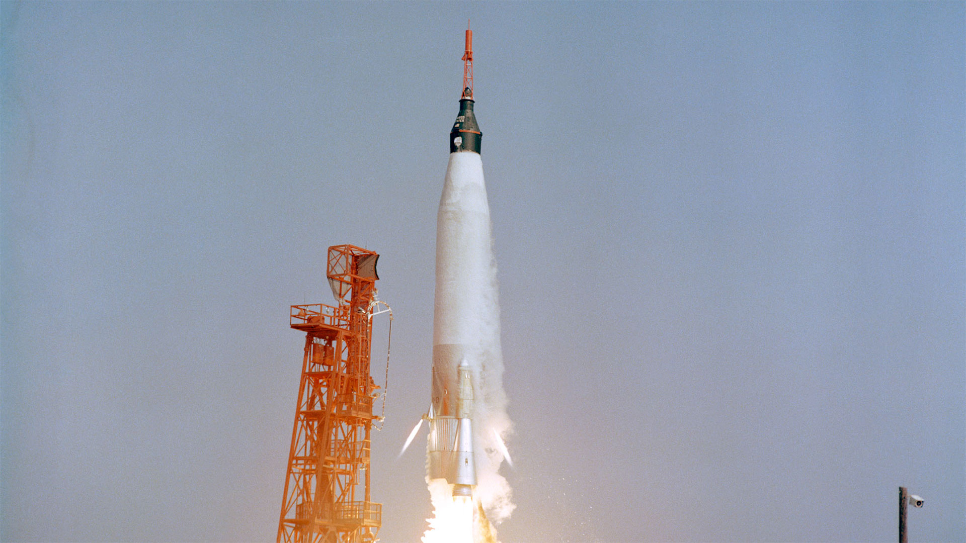 a white rocket topped by a black capsule and a red tower lifts off beside a red support structure
