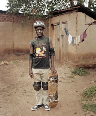 An African man wearing elbow pads, knee pads, a helmet and holding a skateboard.