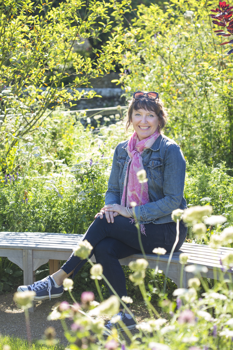 A woman sits on a curved bench in a garden wearing a denim jacket.