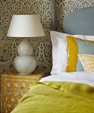 A bedroom with patterned wallpaper, a yellow patterned bedside table with a grey ceramic lamp with white lampshade, and large bed with duck egg blue headboard with bedpost, white bedding, a chartreuse throw, and yellow and blue cushions