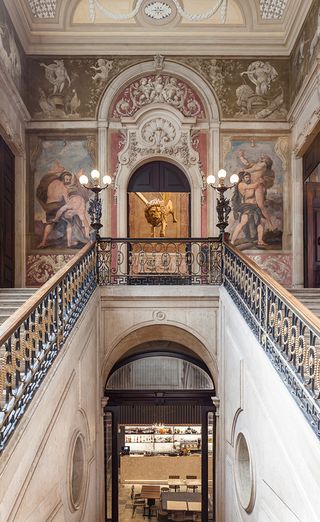 Staircase in the Palacio Chiado. Beautiful iron fence, with light fixtures at the top. The walls are decorated with elaborate frescoes and stained glass windows. We see a restaurant area below the staircase.