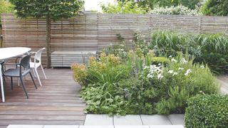 A garden scene with wooden fence and decking and large garden bed with a lovely outdoor dining table