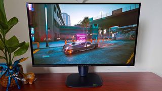 Alienware AW2725Q gaming monitor with Cyberpunk 2077 gameplay on screen featuring car on road in night city and neon lights in backdrop.