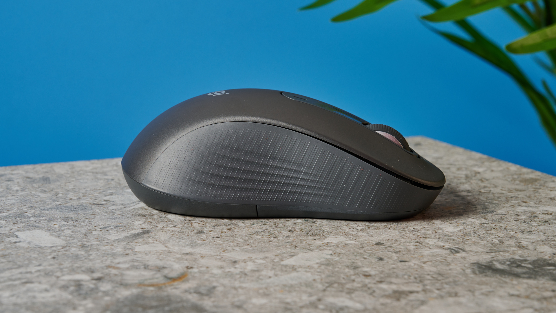 a black mouse with a scroll wheel, two side buttons, and grooved edges, with a logi logo, sits atop a pale table with a blue background