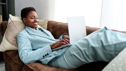 A young woman smiles as she types on her laptop while lying on the sofa.
