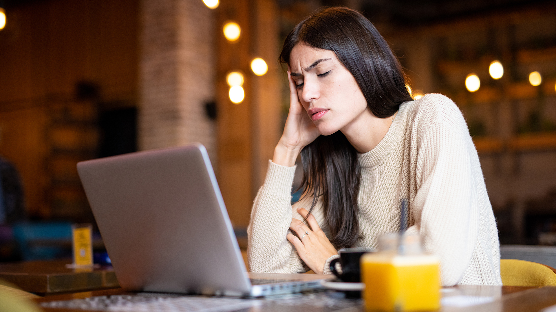 A woman sits in a caf&amp;eacute; with an open laptop, tired overworked, with headache, head leaned on hand.