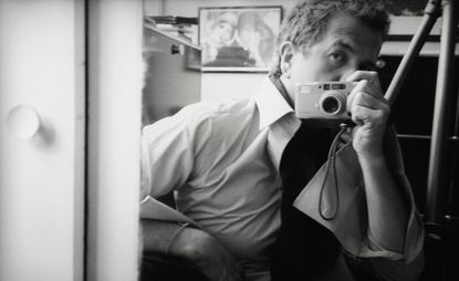 Black and white self portrait image of Mario Testino, sat looking in a mirror, camera in hand taking his own picture
