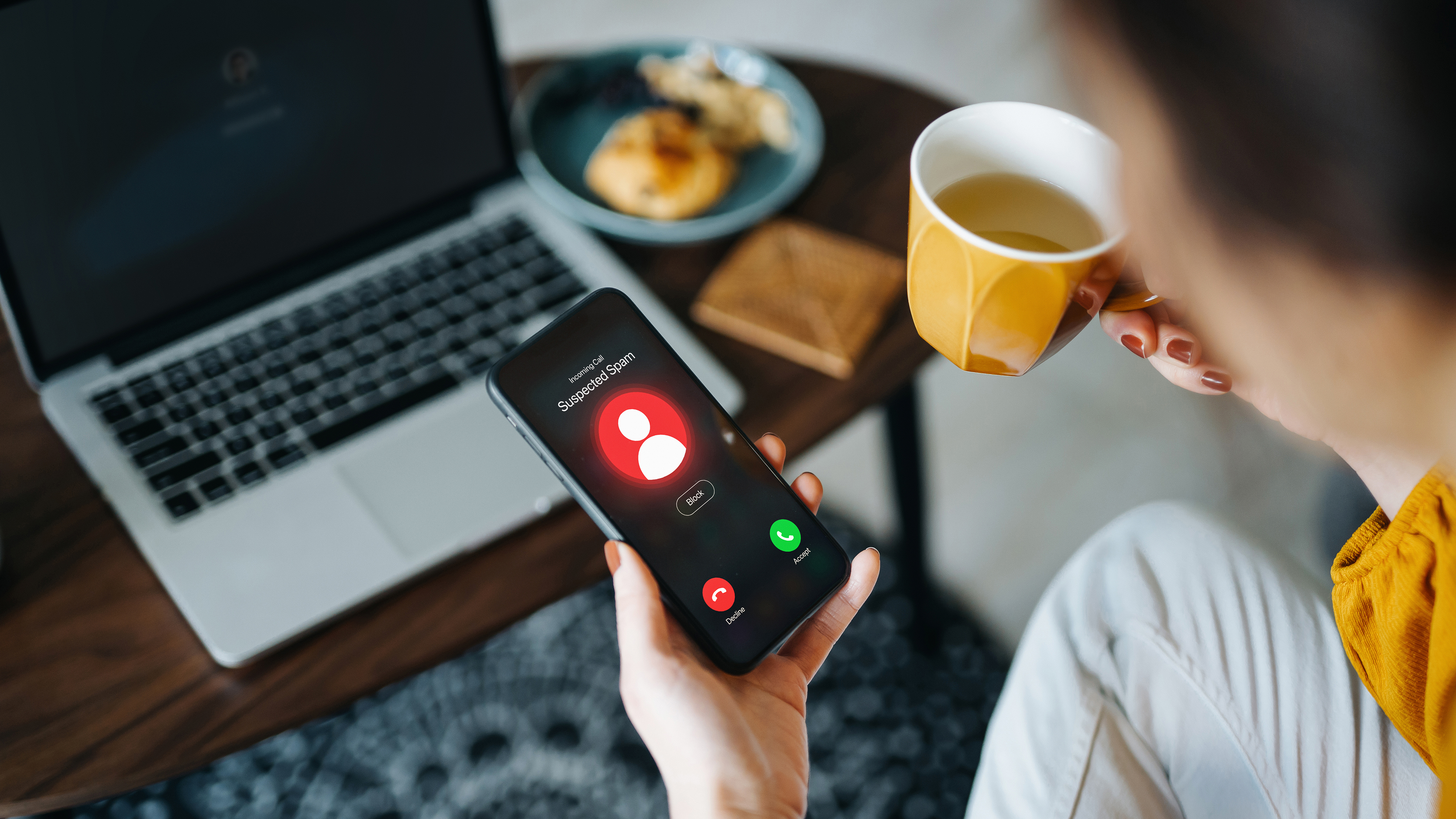 A hand holding a phone showing a suspected scam call, next to a hand holding a mug