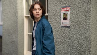 Pip (EMMA MYERS) outside a white building with a missing poster behind her as she looks serious