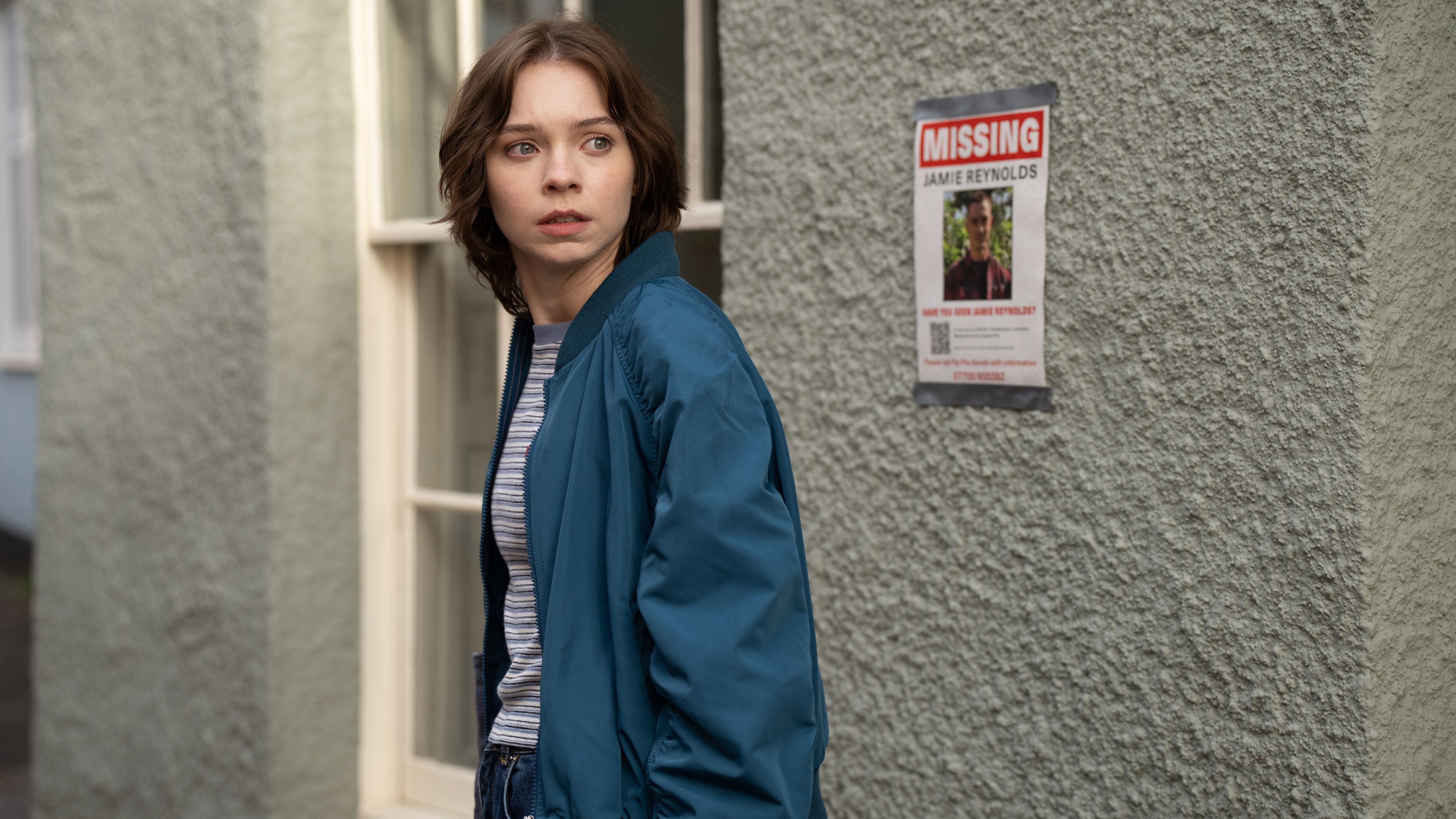 Pip (EMMA MYERS) outside a white building with a missing poster behind her as she looks serious 