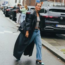 Woman wears black trench coat, black handbag, light wash jeans, gray tee, and heels with teal toe details. 