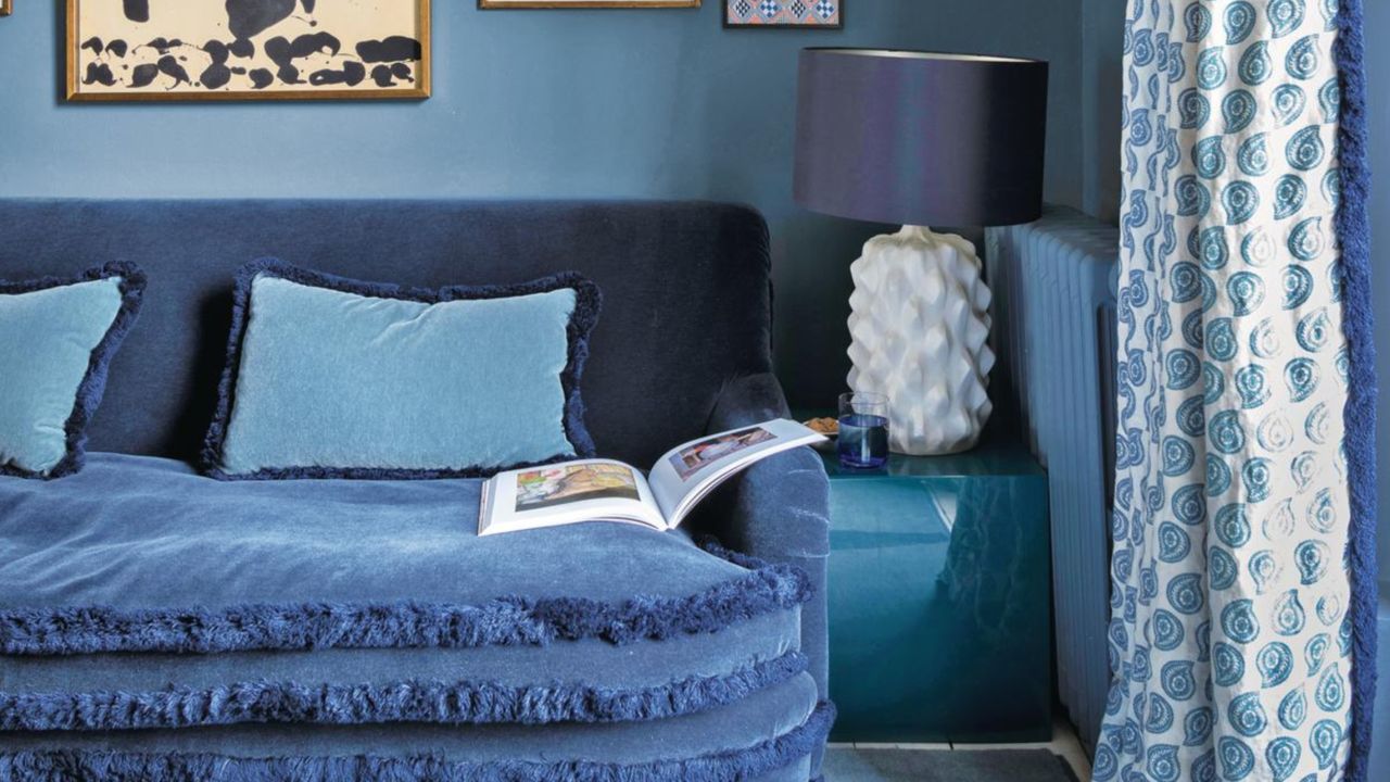 Close-up image of an all-blue living room. The dark blue, velvet couch has tufted trim around the cushions and fringed throw pillows. The blue and white curtain also has tufted blue trim.