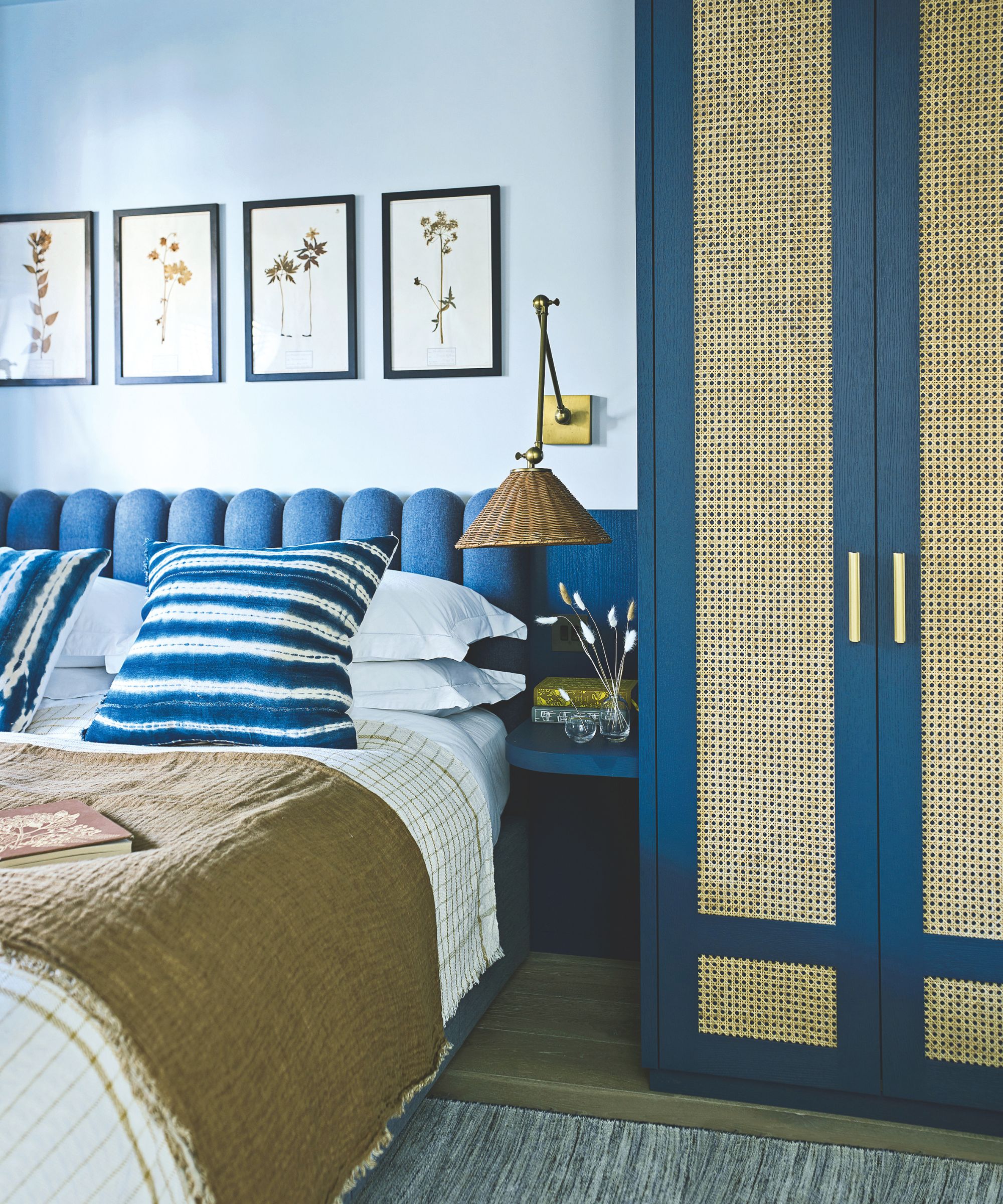 A blue bedroom with blue walls, a large blue bed with an upholstered, scalloped headboard, and a blue closet with rattan panels. Above the bed are four botanical prints in a row in black frames.