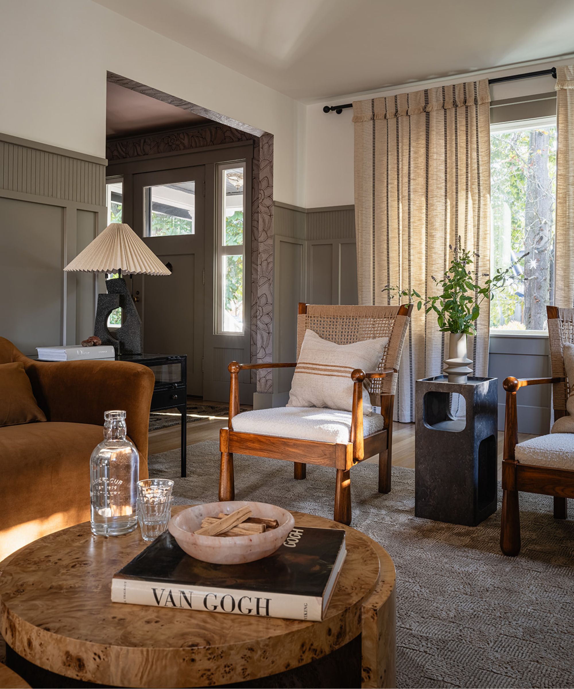 neutral and sage green paneled living room with a round burl wood coffee table, toffee velvet couch, wood and rattan armchairs