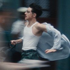 A man (Timothee Chalamet as Marty Mauser) wears a white tank top and carries a black bag, with a loose button-up shirt blowing behind him as he runs. The crowd around him is blurred, in a still from 'Marty Supreme.'