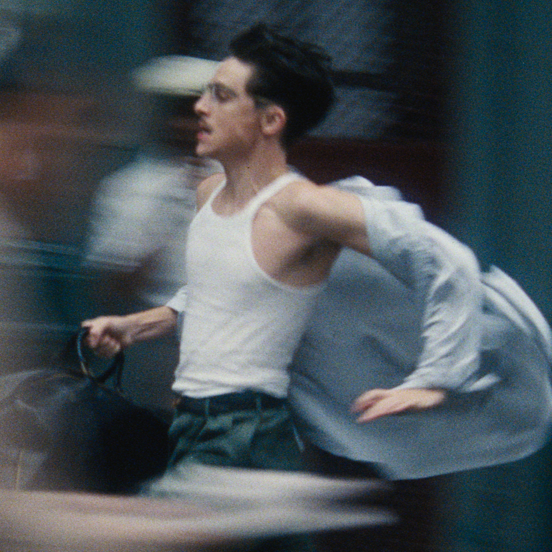 A man (Timothee Chalamet as Marty Mauser) wears a white tank top and carries a black bag, with a loose button-up shirt blowing behind him as he runs. The crowd around him is blurred, in a still from 'Marty Supreme.'