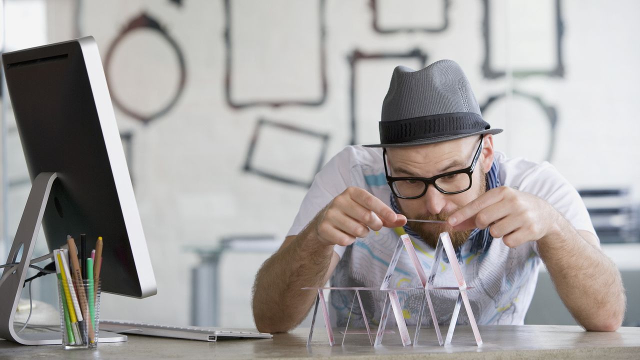 A man wearing a hat builds a house of cards at his desk.