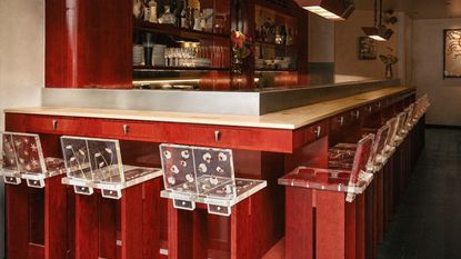 A red-painted wood and chrome bar of a restaurant, decorated with tall, geometrically cut wooden stools with flower-decorated resin backs.
