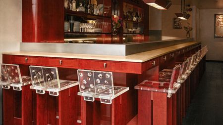 A red-painted wood and chrome bar of a restaurant, decorated with tall, geometrically cut wooden stools with flower-decorated resin backs.